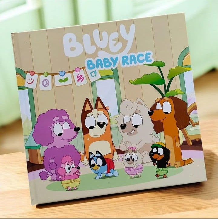 A children's book titled 'Bluey Baby Race' standing upright on a wooden surface. The cover features colorful cartoon dog characters inspired by Bluey, including a purple poodle, an orange dog, a white dog, a brown dog, a blue bird, a pink baby bird, and a black duckling, set against a light wood-paneled background.