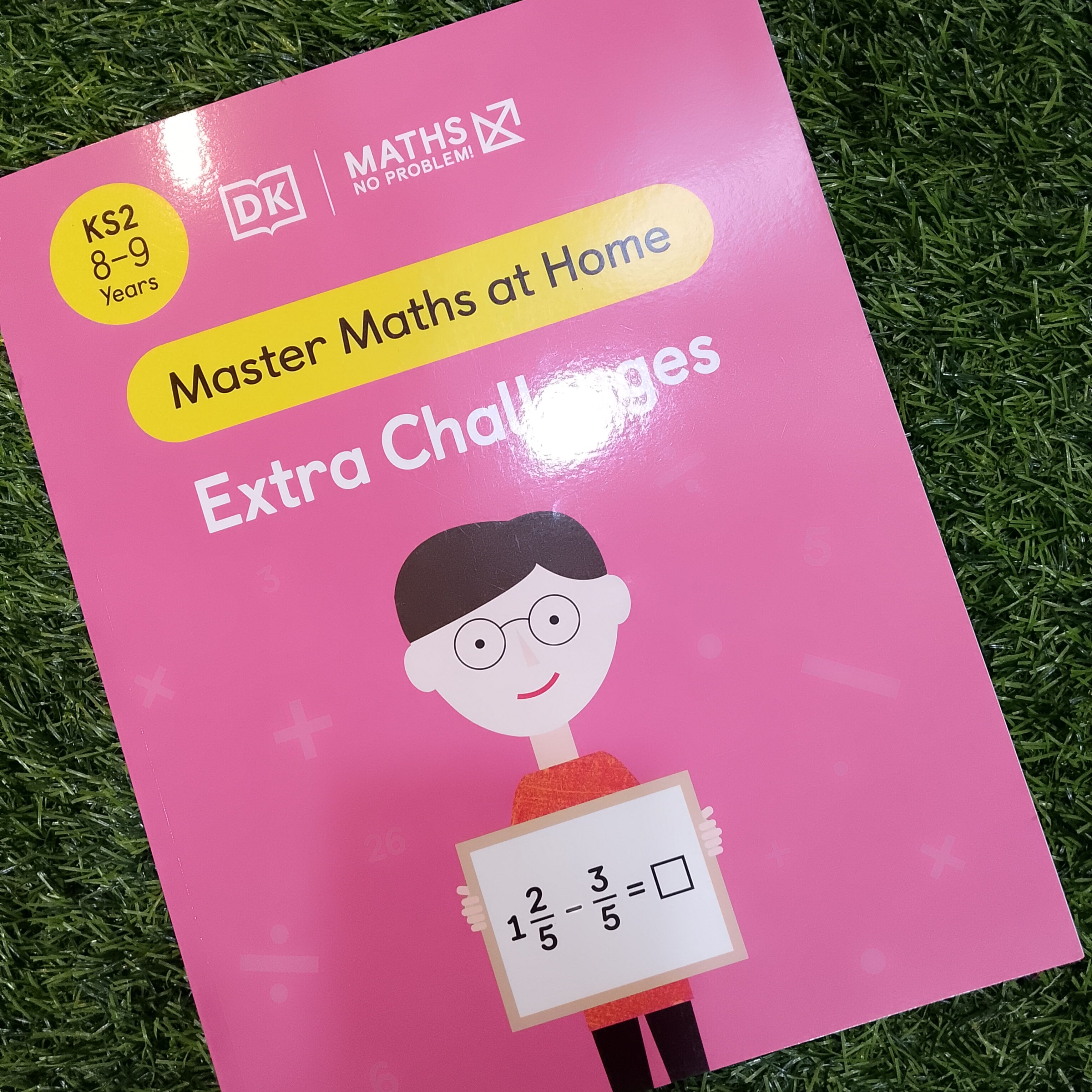Pink book titled 'Master Maths at Home Extra Challenges' on a grass background