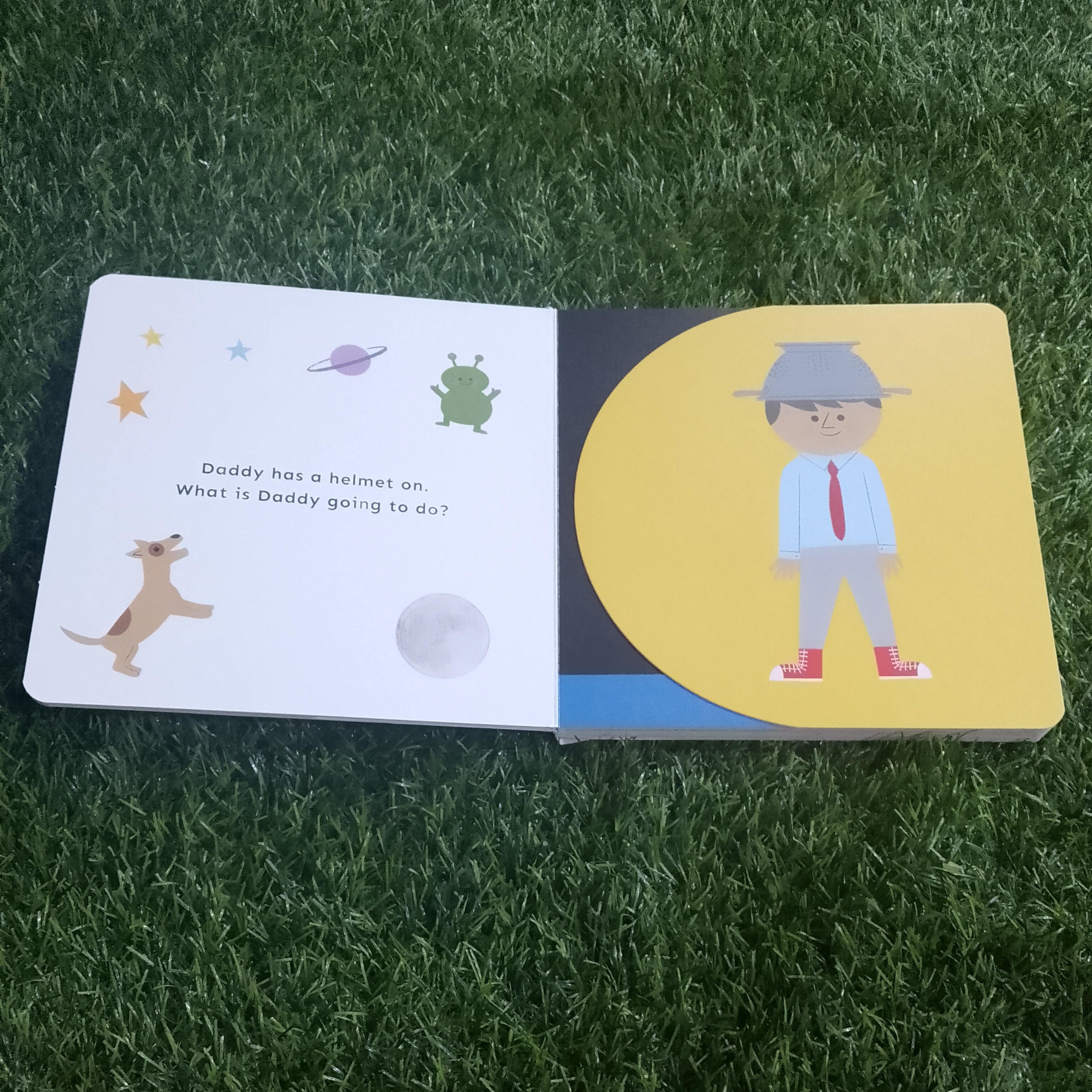 Children's book open on grass with illustrations of a dog, frog, and person wearing a hat and tie.