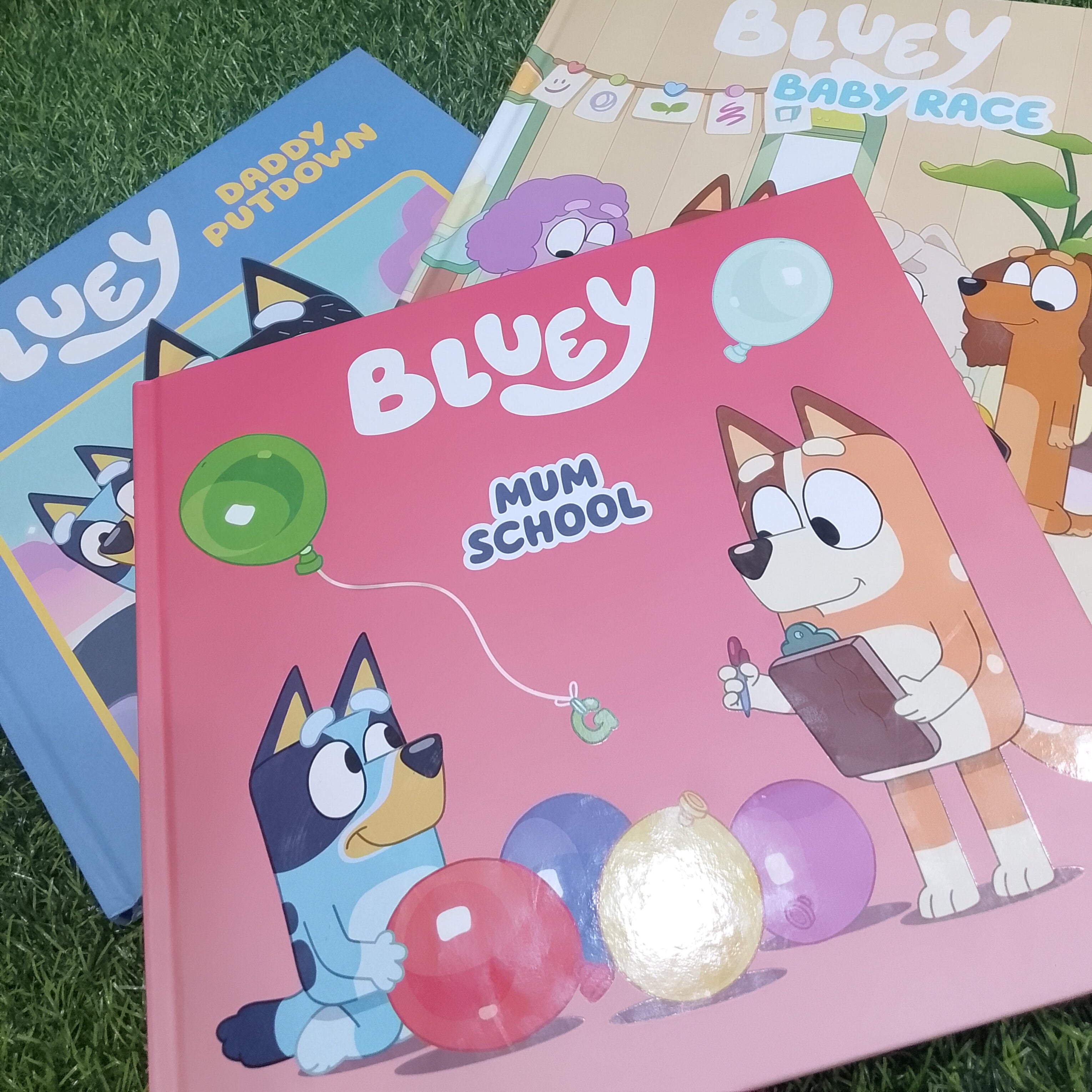 Three children's Bluey-themed books fanned out on artificial green grass. The front book is pink with the title 'Bluey Mum School' and shows Bluey and Bingo with balloons.